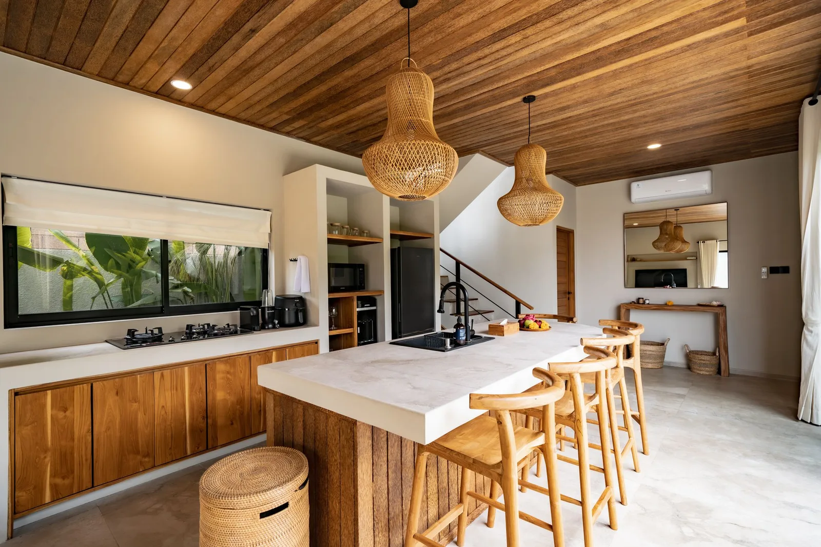 A bright kitchen features a large white island, wooden bar stools, built-in cabinetry, and woven pendant lights.