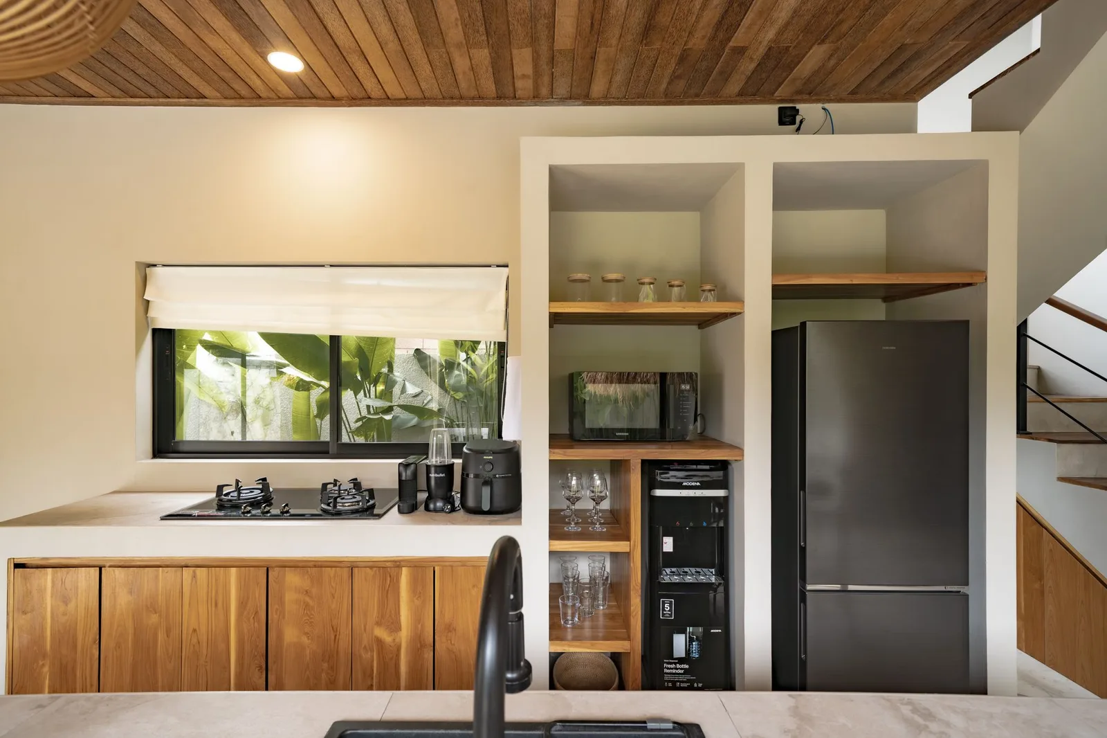 A modern kitchen with wood cabinetry, a wide window, open shelving, and a stainless-steel refrigerator.