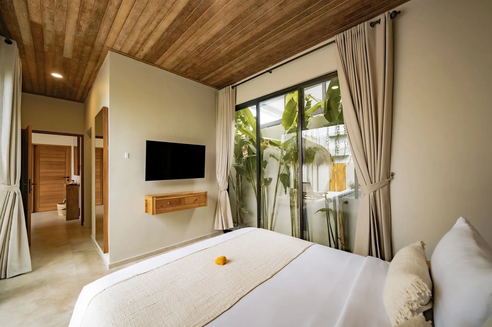 A bedroom with a wall-mounted television, wooden ceiling, and glass doors opening onto a leafy patio.