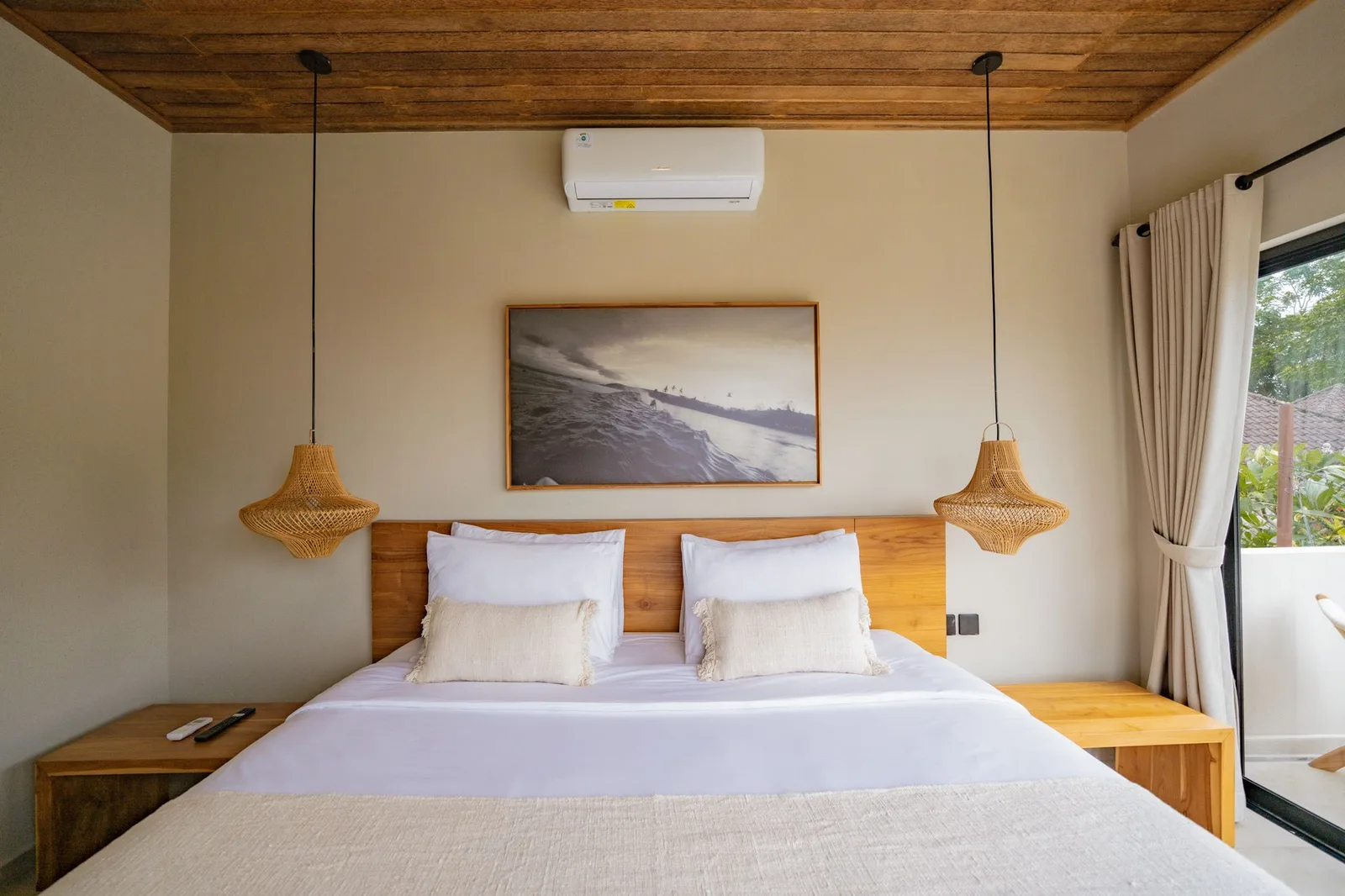 A bedroom with a neatly made bed, wooden headboard, pendant lights, and coastal artwork above it.