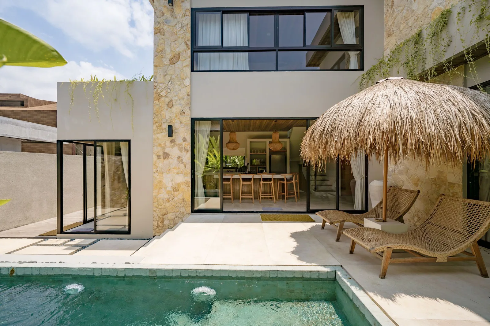 A modern stone-clad house opens onto a patio with a small pool, woven chairs, and a thatched umbrella.
