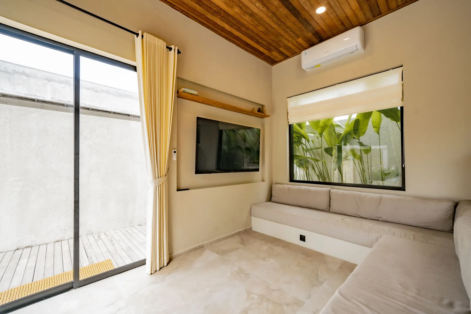 A bright sitting area features a built-in sofa, wall-mounted television, large windows, and a wood-paneled ceiling.
