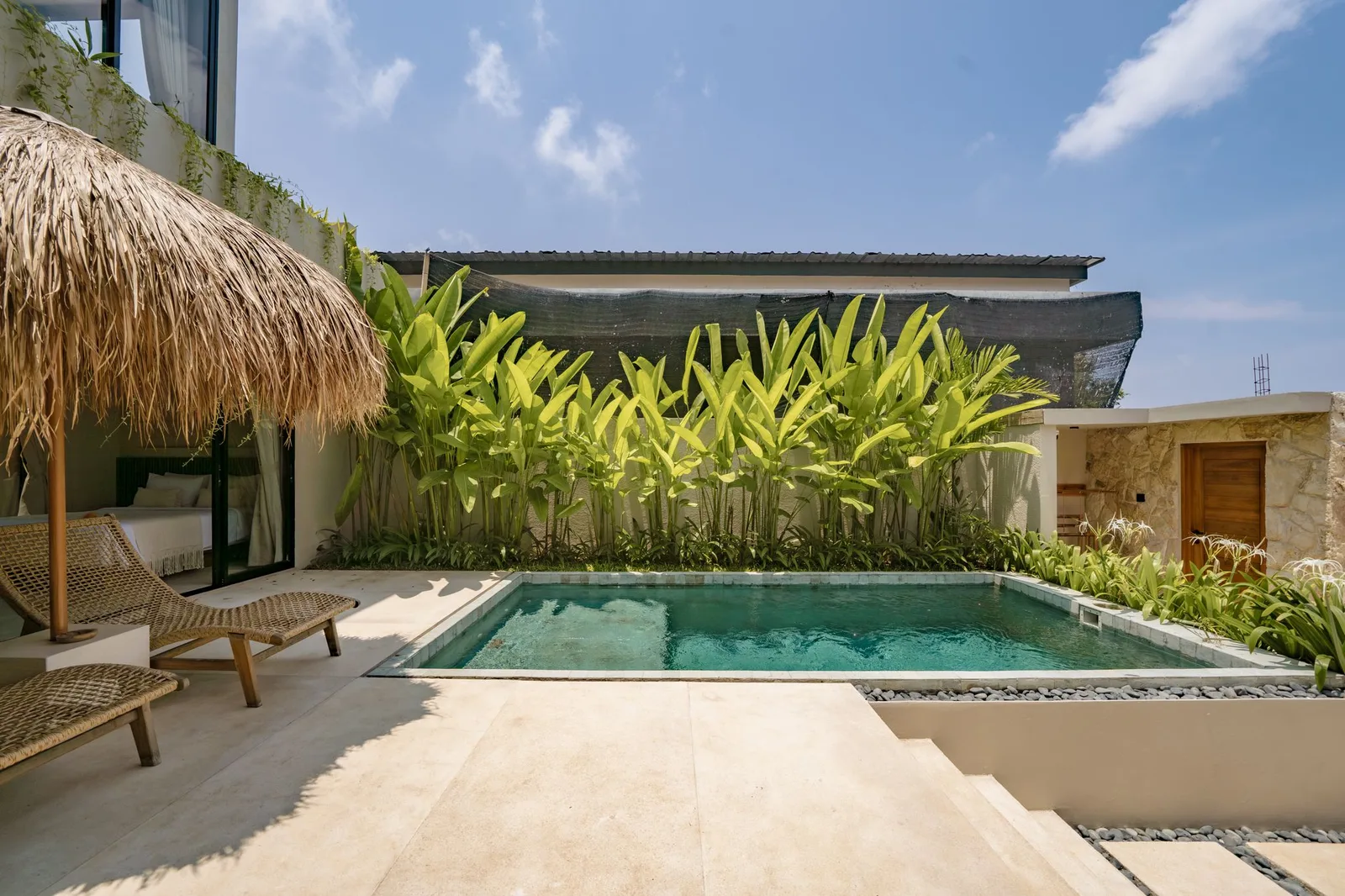 A small outdoor pool sits beside a thatched shade structure, tropical plants, and a pale stone terrace.
