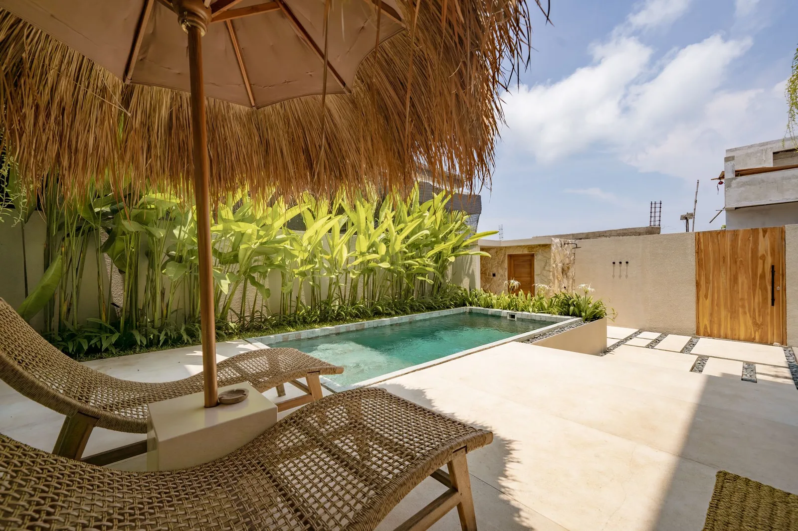 A small outdoor pool sits beside woven lounge chairs, leafy plants, and a thatched shade canopy.