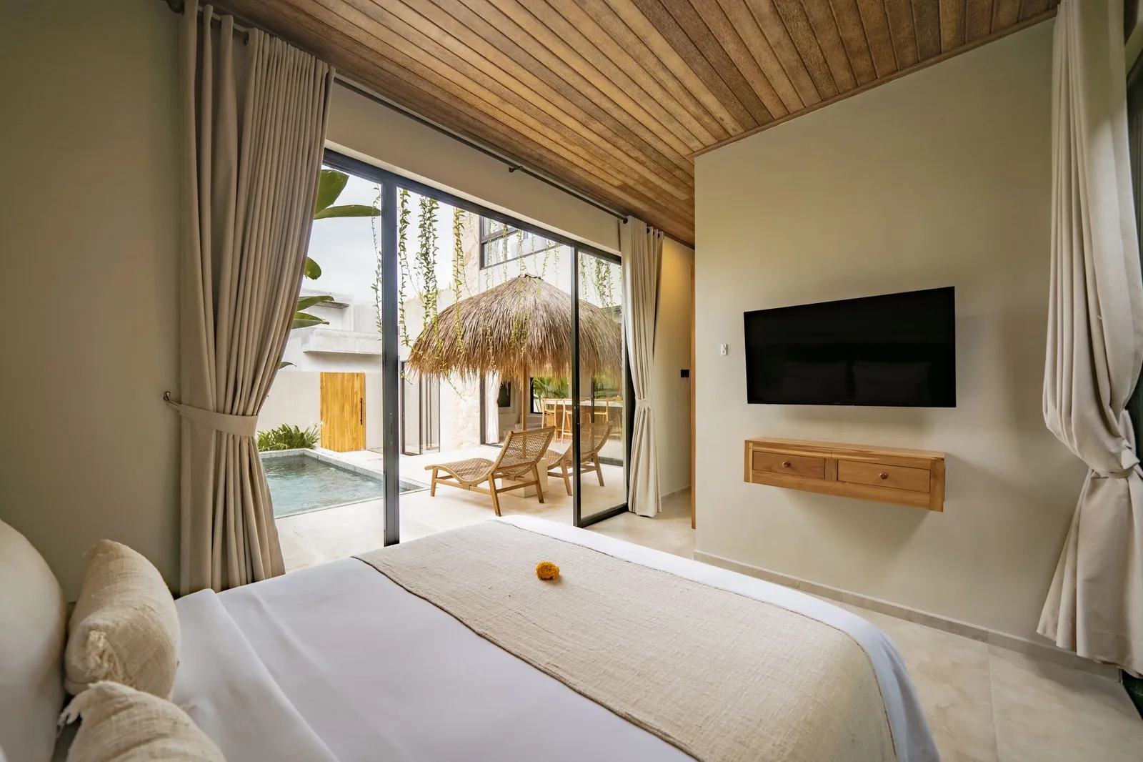 A bright bedroom with a wooden ceiling, wall-mounted television, and sliding doors opening to a poolside patio.