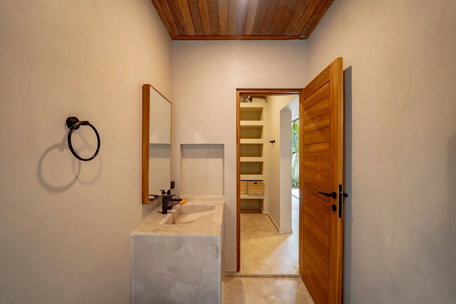 A bathroom vanity with a rectangular sink sits beside an open wooden door and recessed shelves.