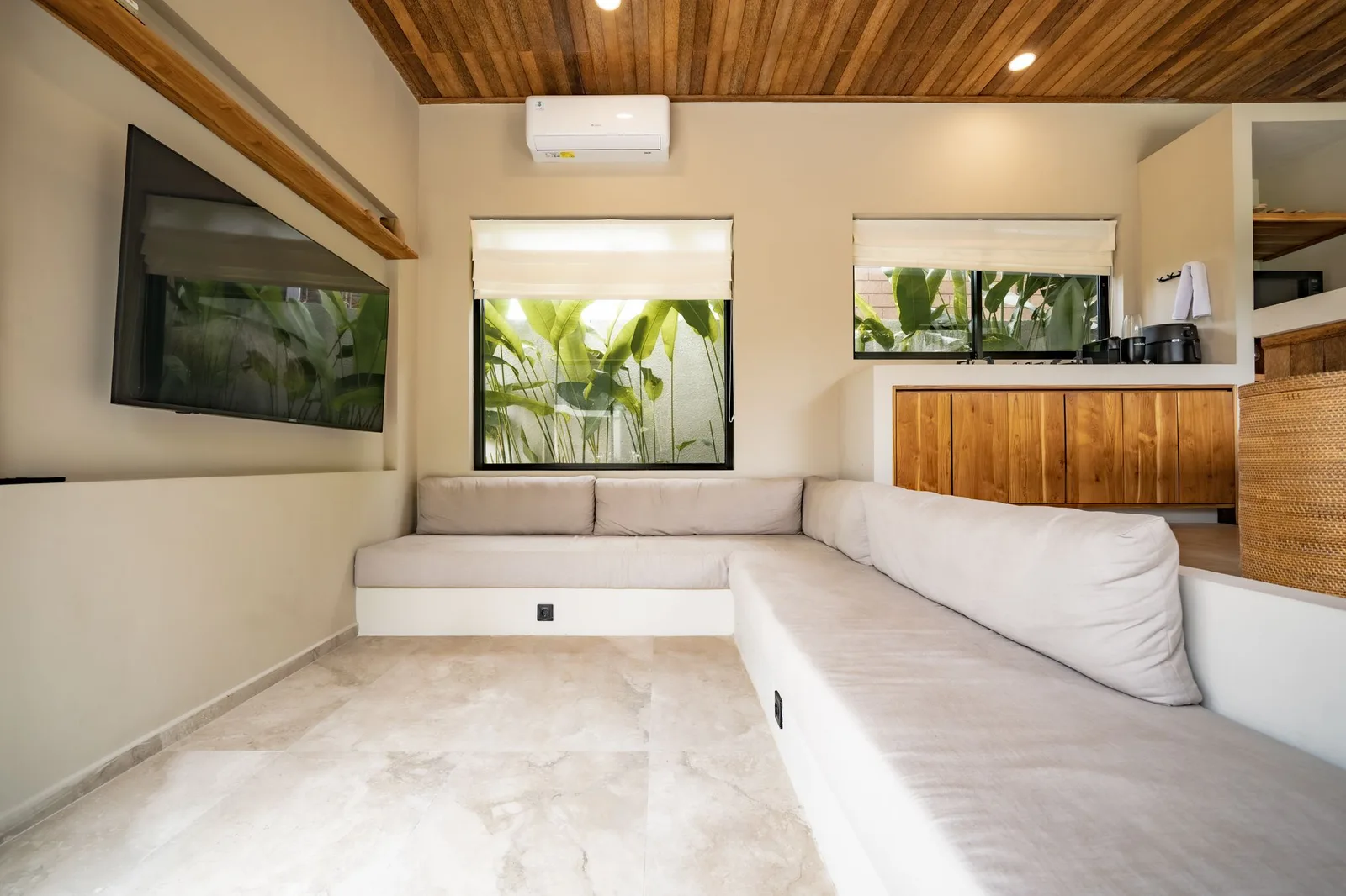 A bright living room has an L-shaped white sofa, leafy windows, and a wood-paneled ceiling.