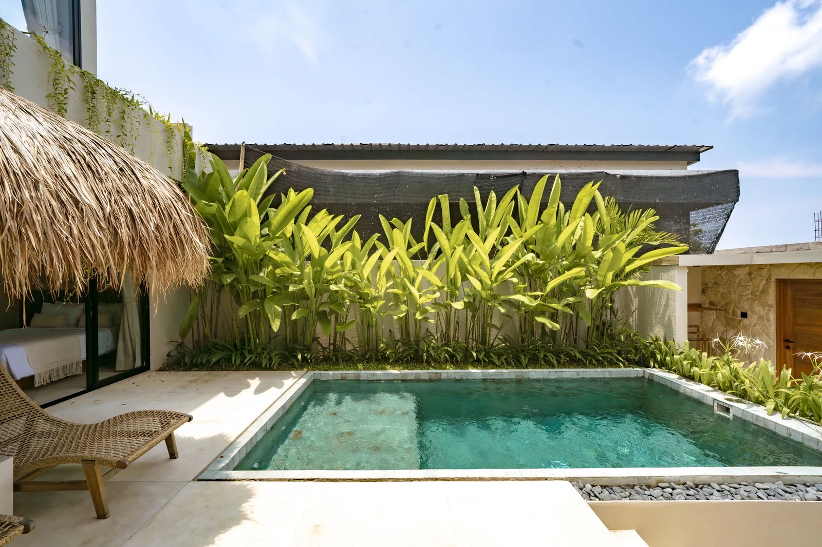 A turquoise swimming pool sits beside a thatched shade canopy and a dense row of tropical plants.
