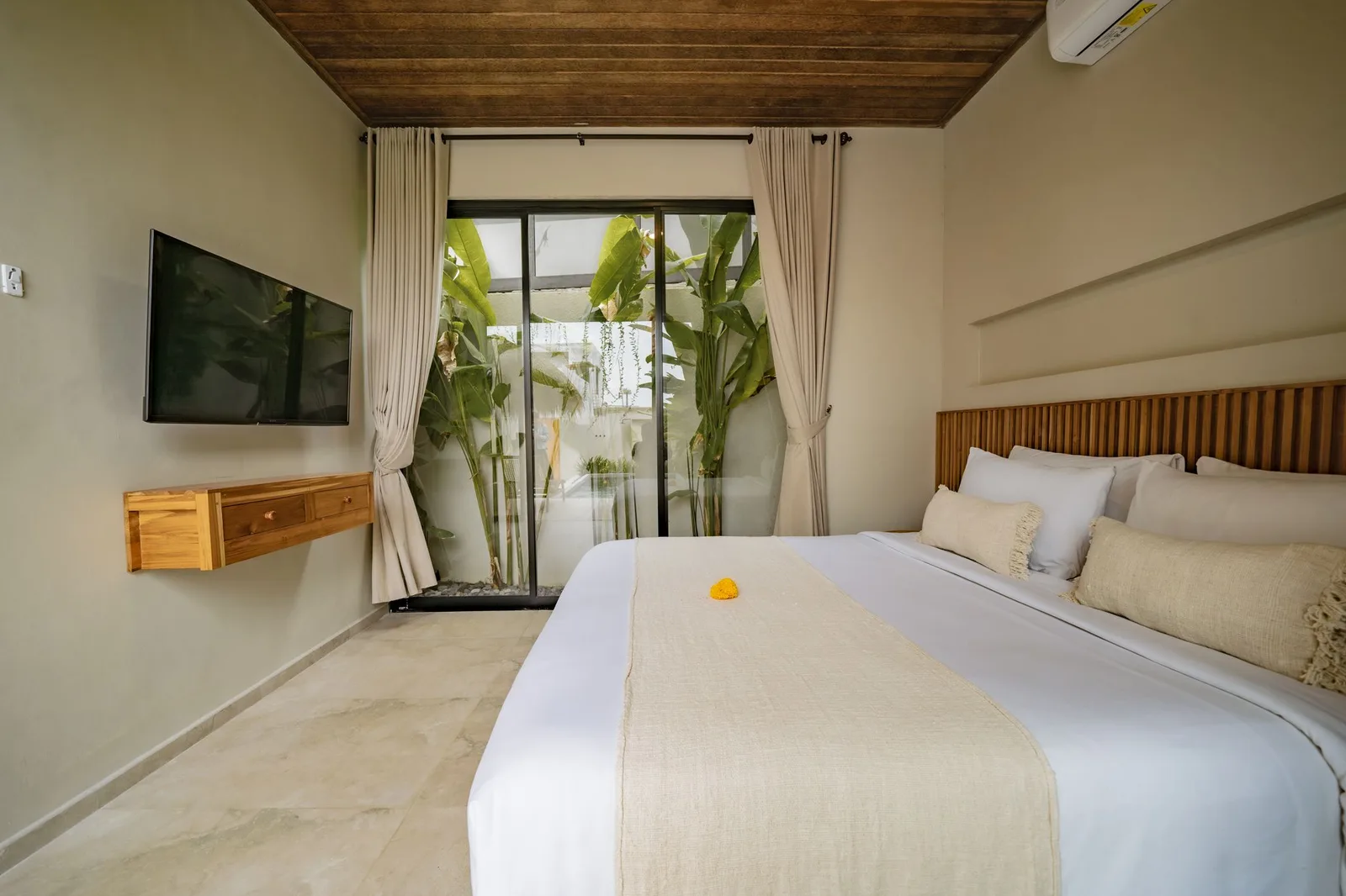 A bedroom with a king bed, wooden headboard, wall-mounted television, and sliding glass doors opening to greenery.