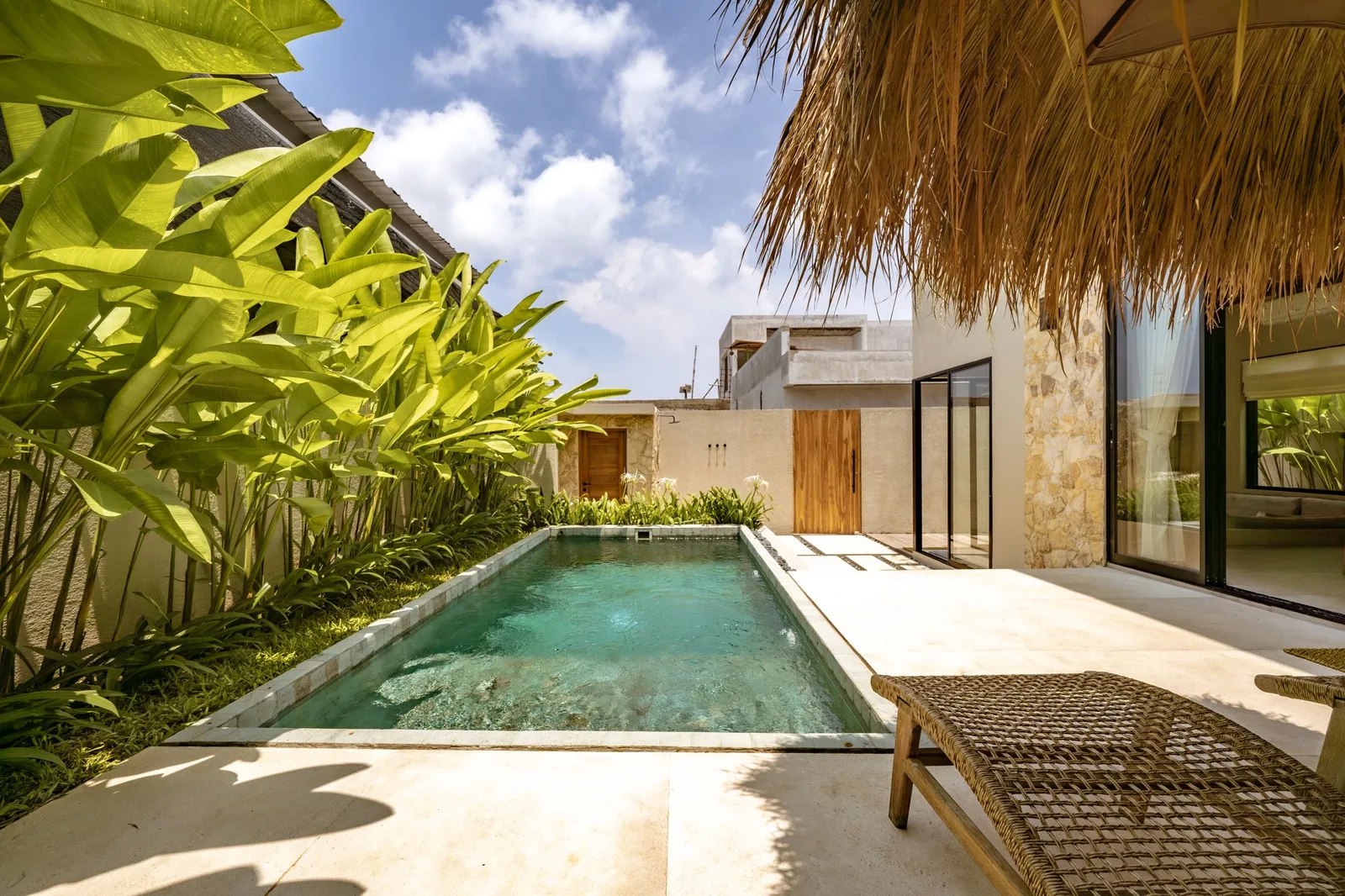 A small outdoor pool sits beside a modern villa, with tropical plants and a thatched overhang.