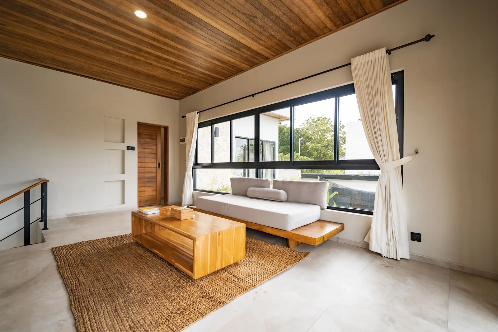 A bright living room with a low white sofa, wooden coffee table, woven rug, and large windows.