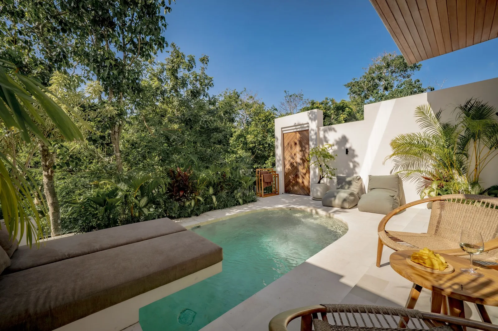 Outdoor pool patio with cushioned loungers, a woven chair, and tropical plants beside a white wall.