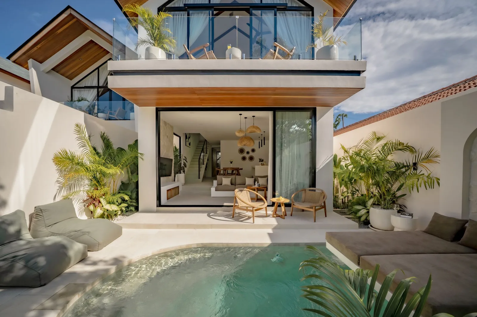 A modern house opens onto a small pool, with a furnished patio, tropical plants, and an upper balcony.
