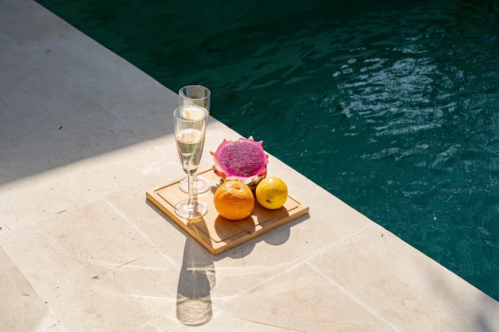 Poolside scene with a wooden tray holding two drinks, oranges, and a halved dragon fruit.