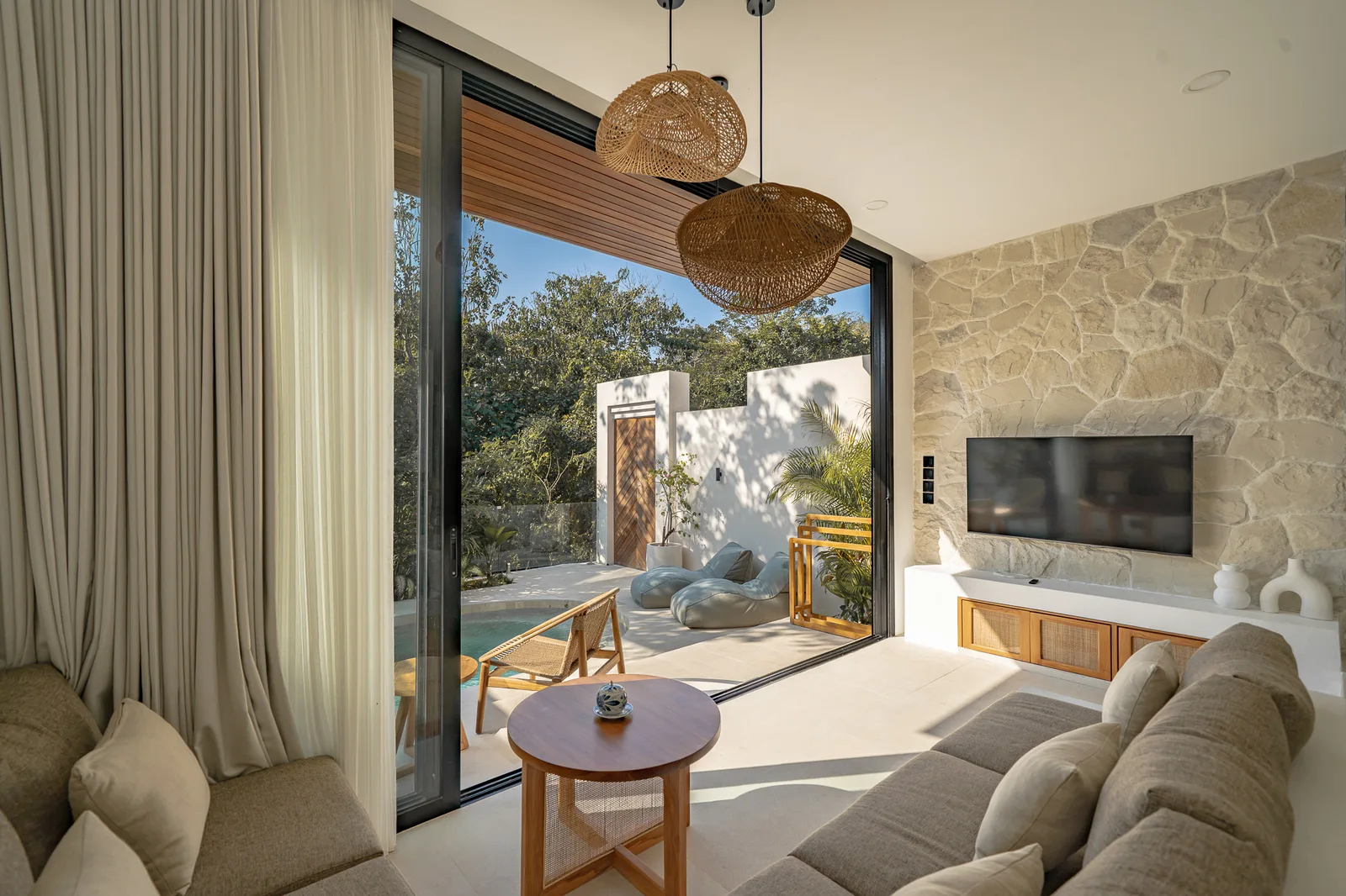 A bright living room opens onto a patio, with a stone feature wall, television, and woven pendant lights.