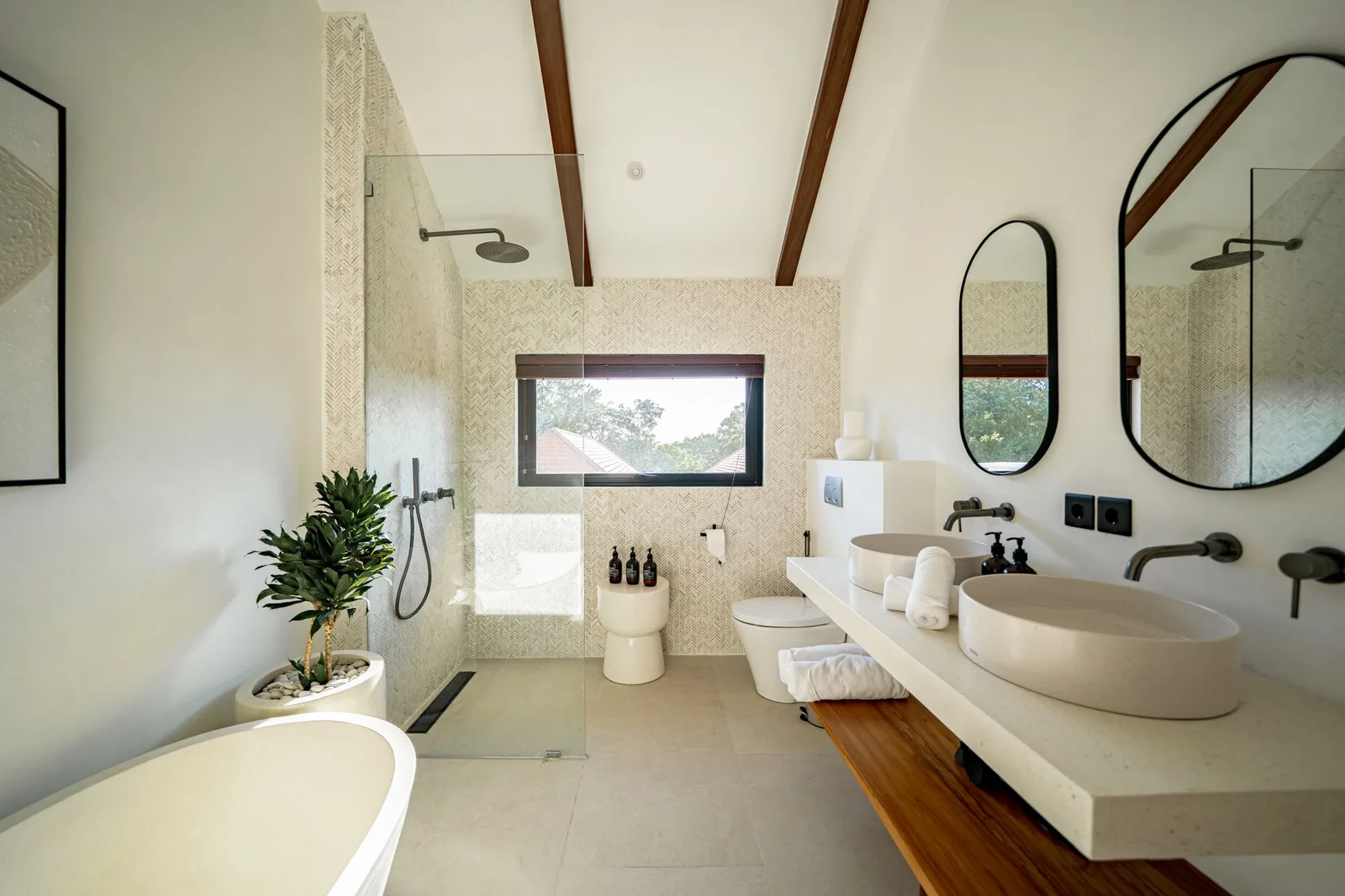 A bright bathroom features a freestanding tub, walk-in shower, double sinks, and a window overlooking greenery.
