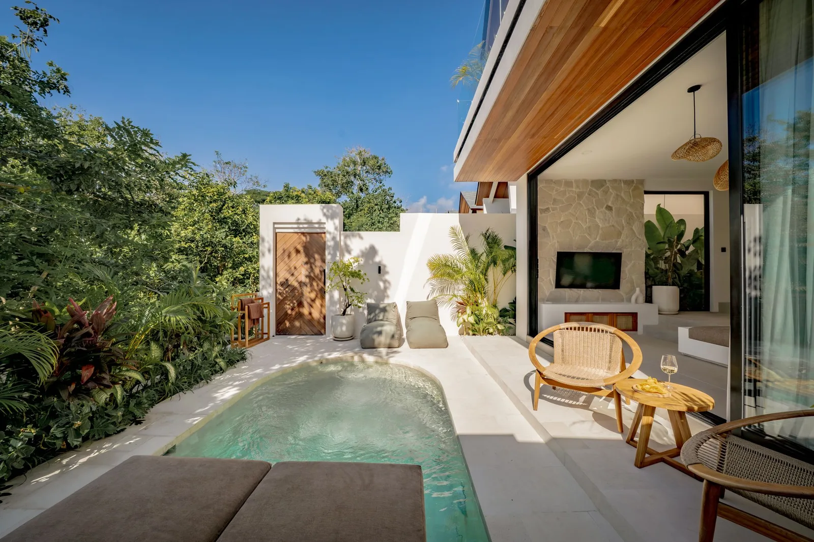 A small outdoor pool sits beside a modern house, with lounge cushions, wicker chairs, and lush plants.
