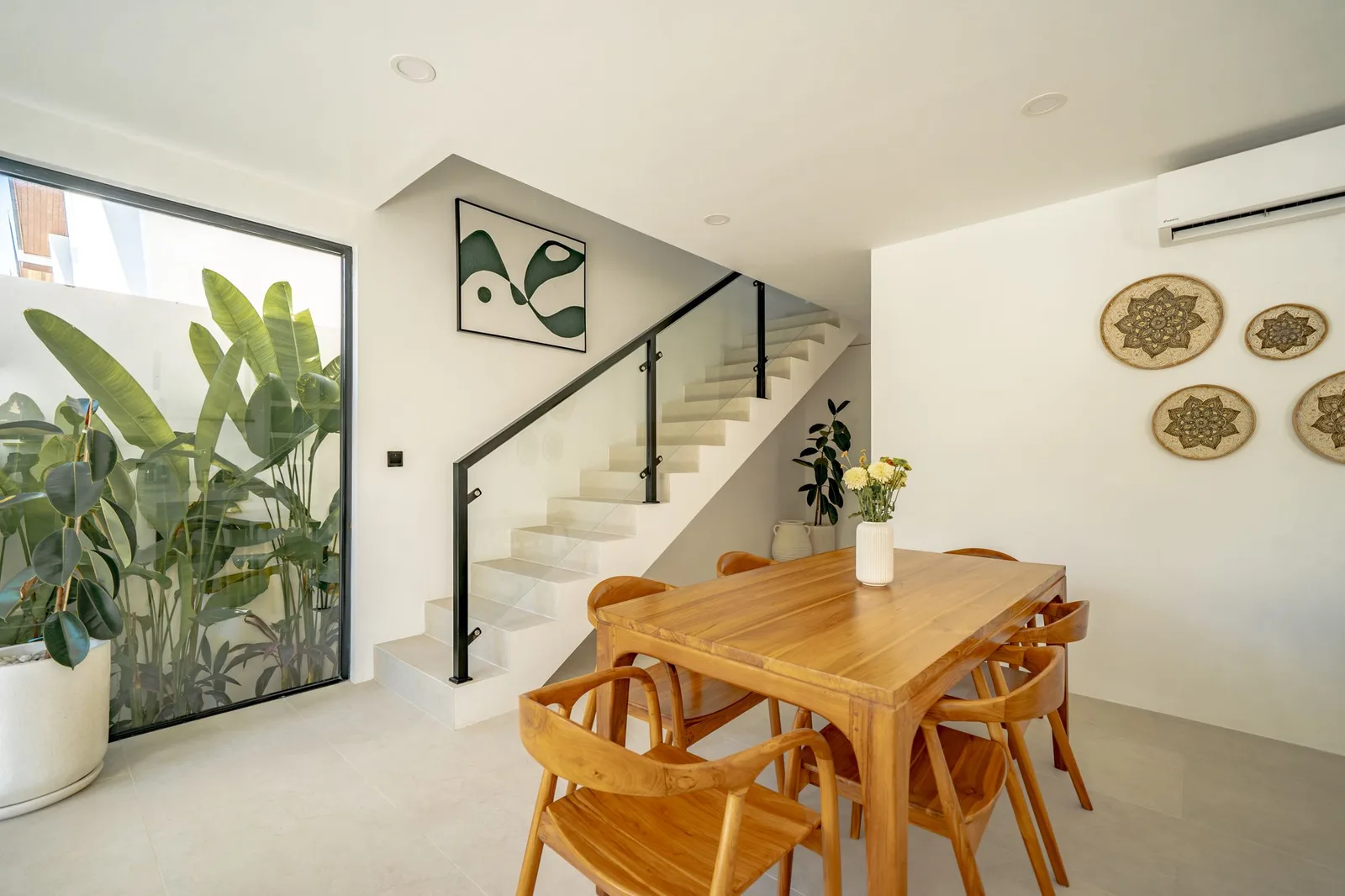 A bright dining area features a wooden table and chairs beside a staircase and large potted plants.