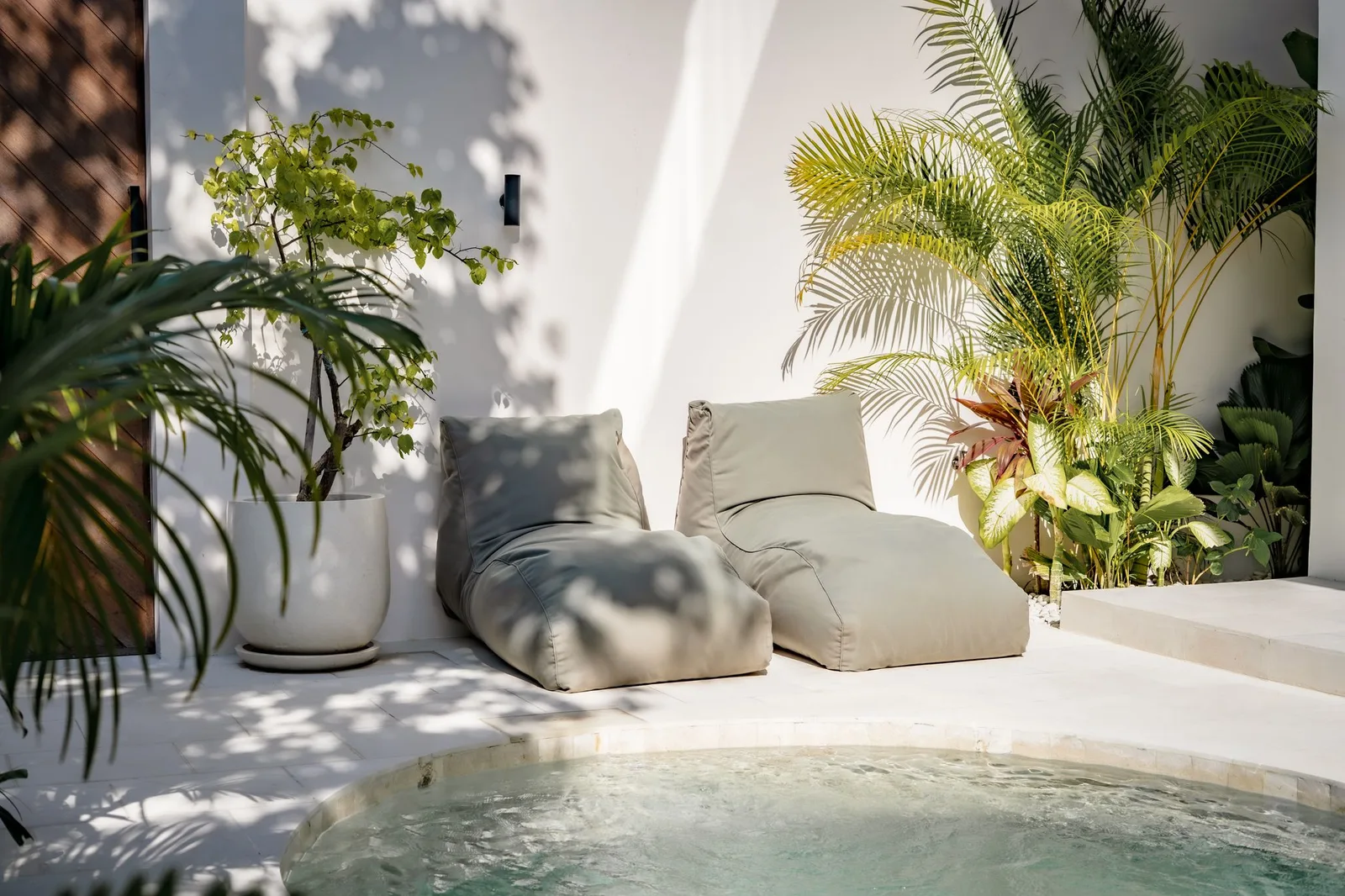 Outdoor poolside lounge with cushioned chairs, potted plants, and a curved pool edge in the foreground.