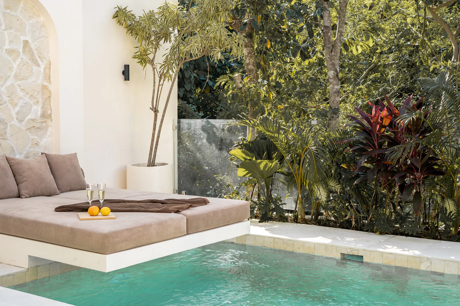 A turquoise pool sits beside a raised cushioned daybed, with leafy tropical plants along the wall.