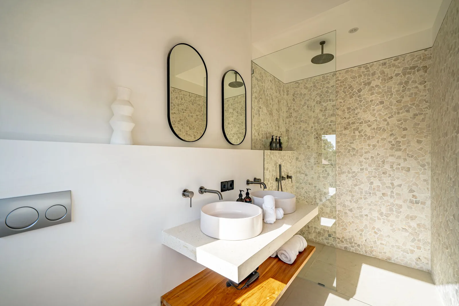 A modern bathroom features a double vanity with vessel sinks, oval mirrors, and a stone-tiled shower wall.