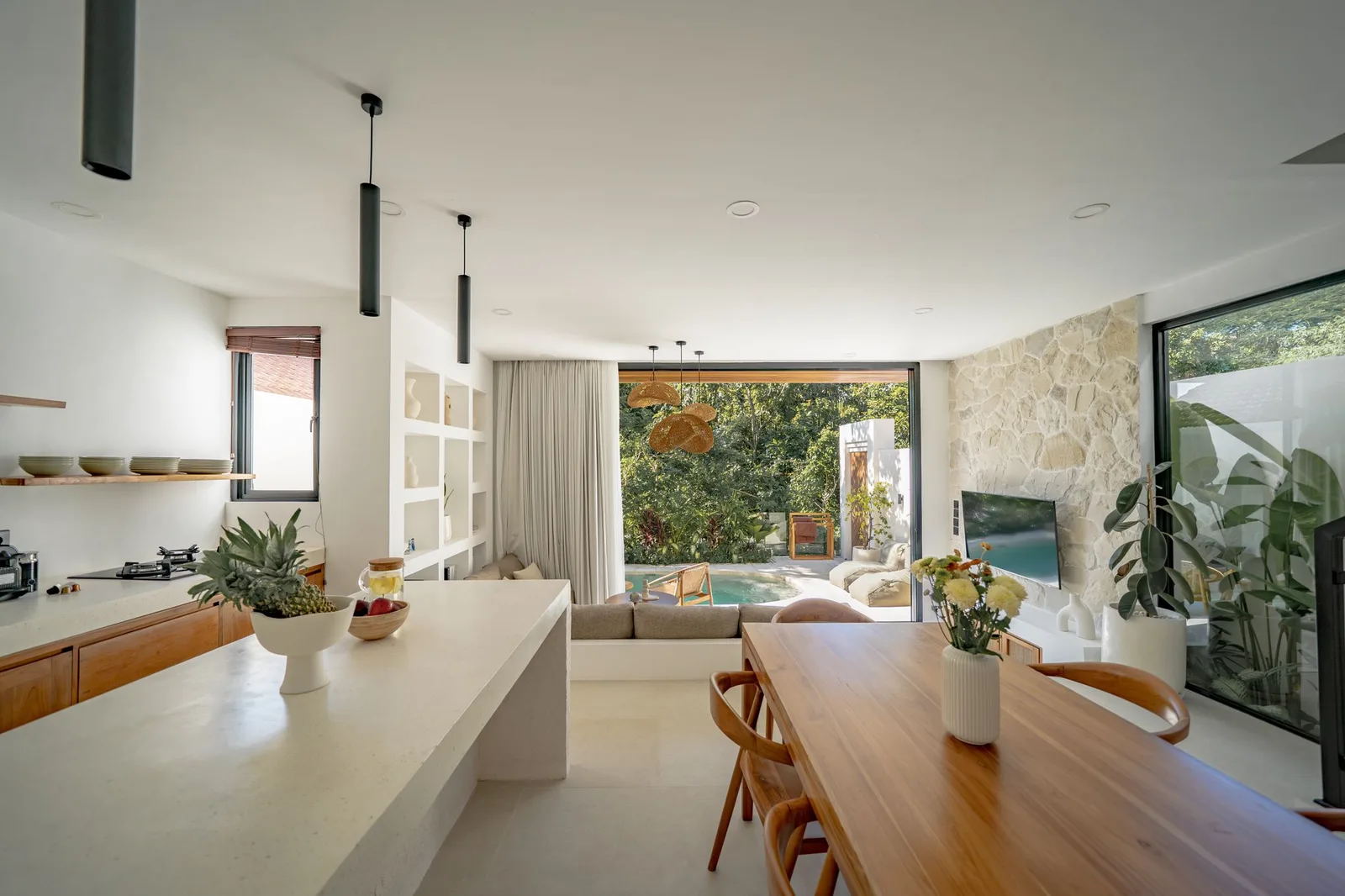 Open-plan kitchen and dining area with a long wooden table, stone wall, and garden-facing windows.