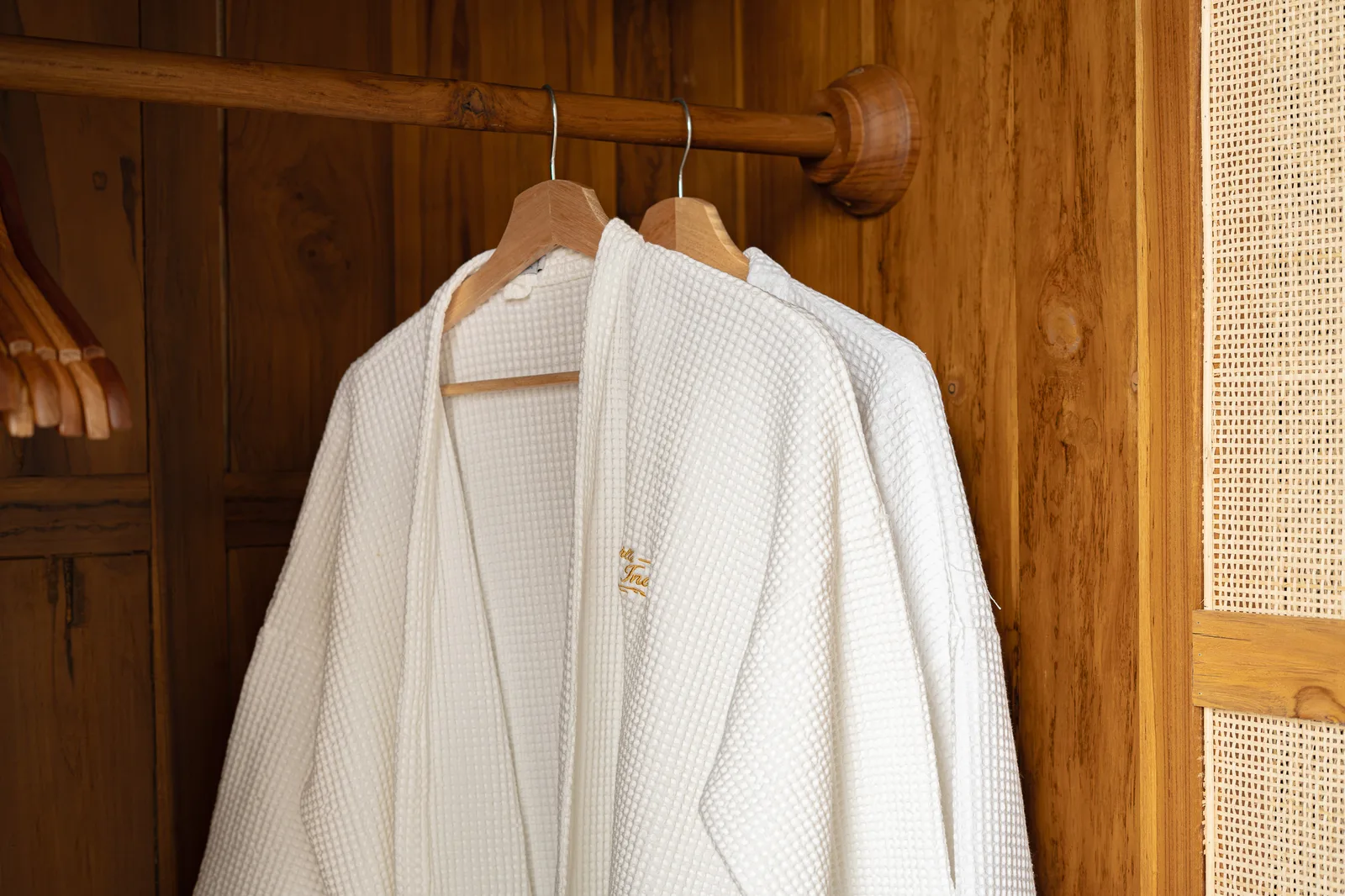 Two white robes hang on wooden hangers inside a wooden wardrobe.