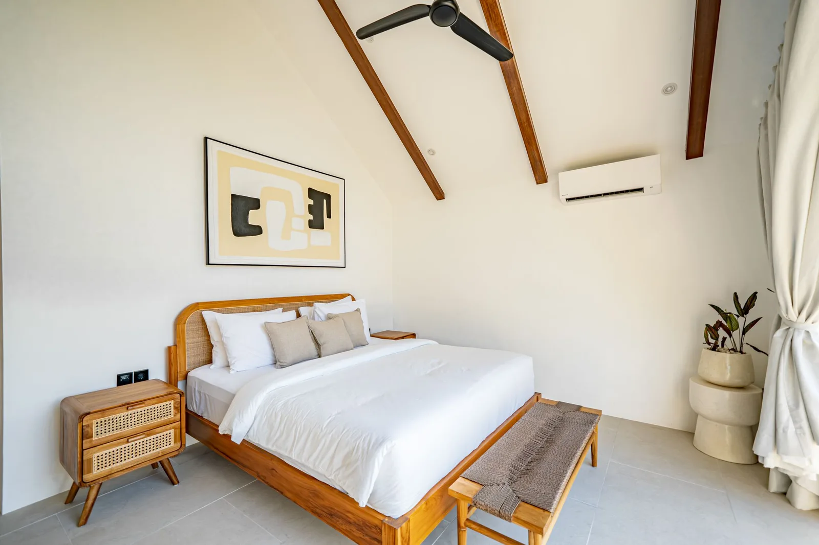 A bright bedroom features a wooden bed with white linens, a bedside table, and exposed ceiling beams.