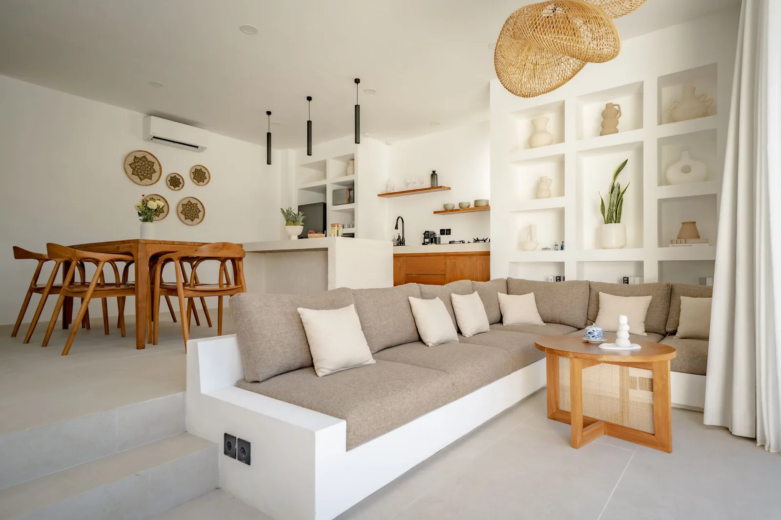 A bright open-plan living and dining area features a built-in sofa, wooden table, and a white kitchen.