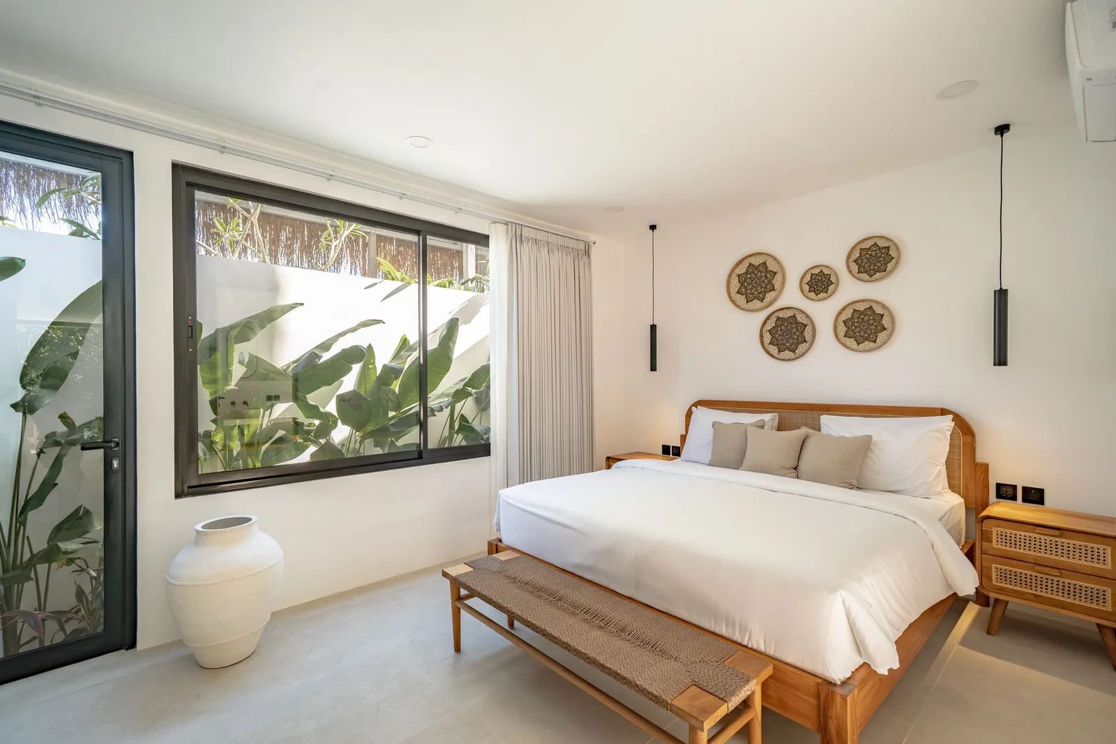 A bright bedroom has a wooden bed with white linens, woven wall decor, and large windows overlooking leafy plants.