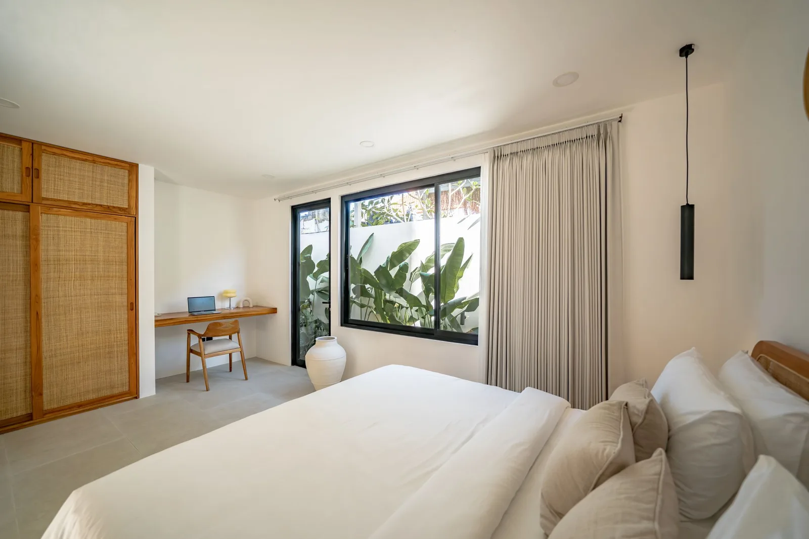 A bright bedroom with a large bed, woven wardrobe doors, and broad windows overlooking leafy plants.