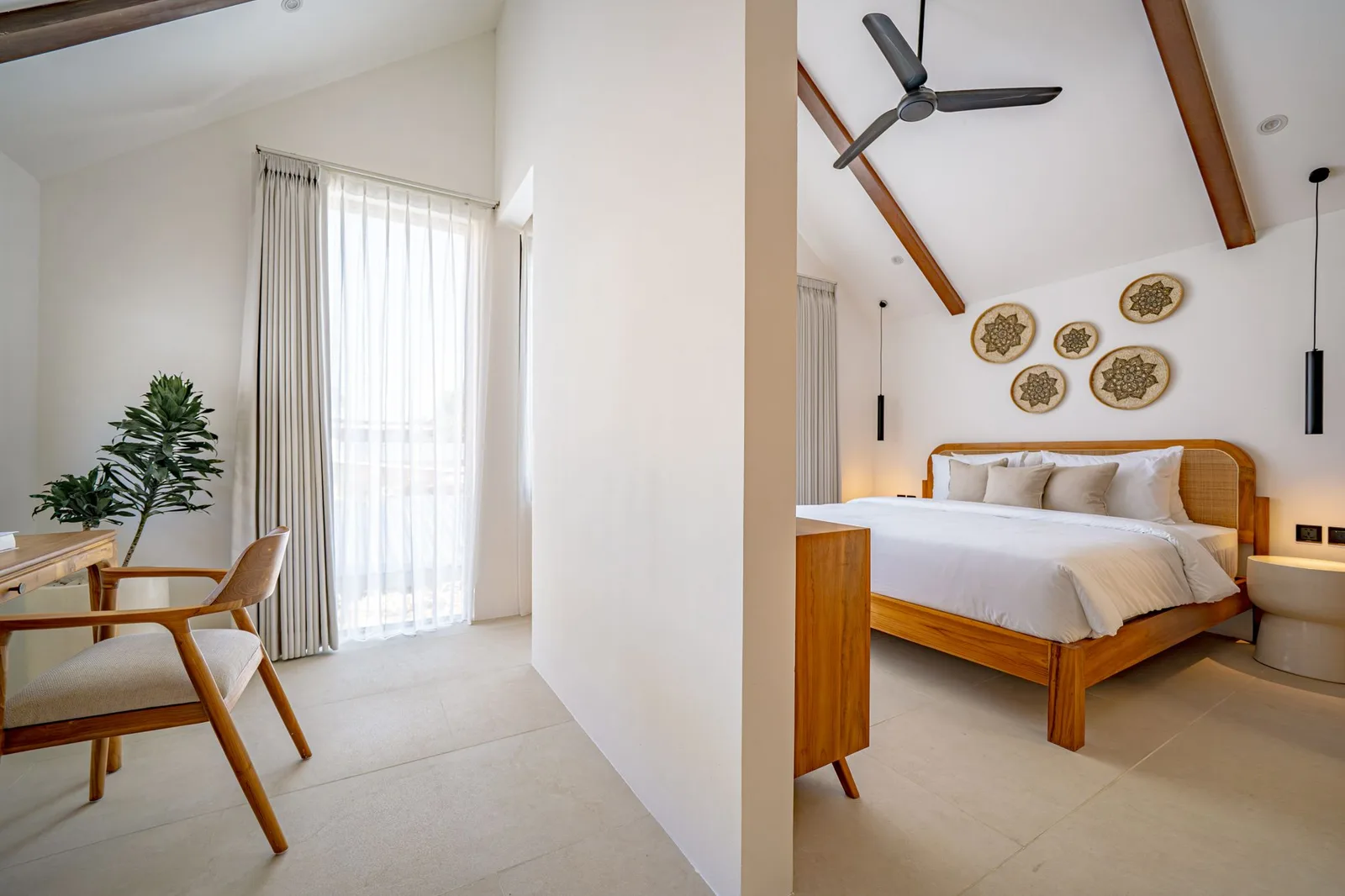 A bright bedroom has a wooden bed, white bedding, a ceiling fan, and a window with sheer curtains.