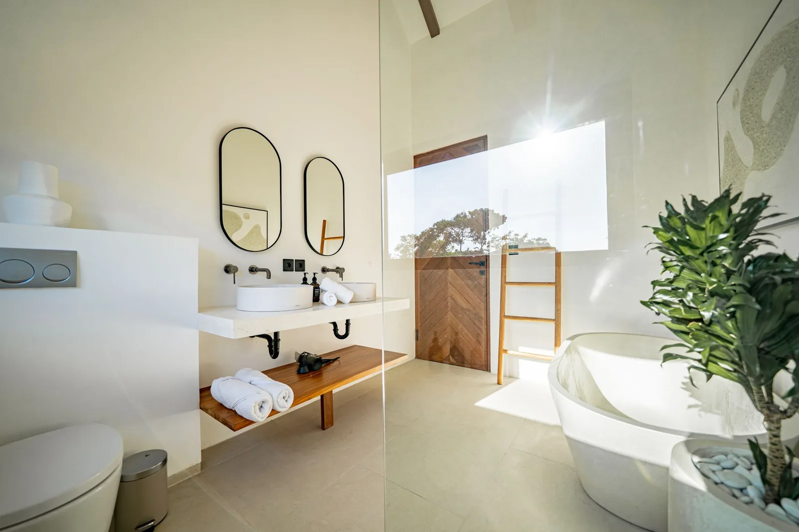 A bright modern bathroom has a white sink, wall mirrors, a wooden towel shelf, and a freestanding bathtub.