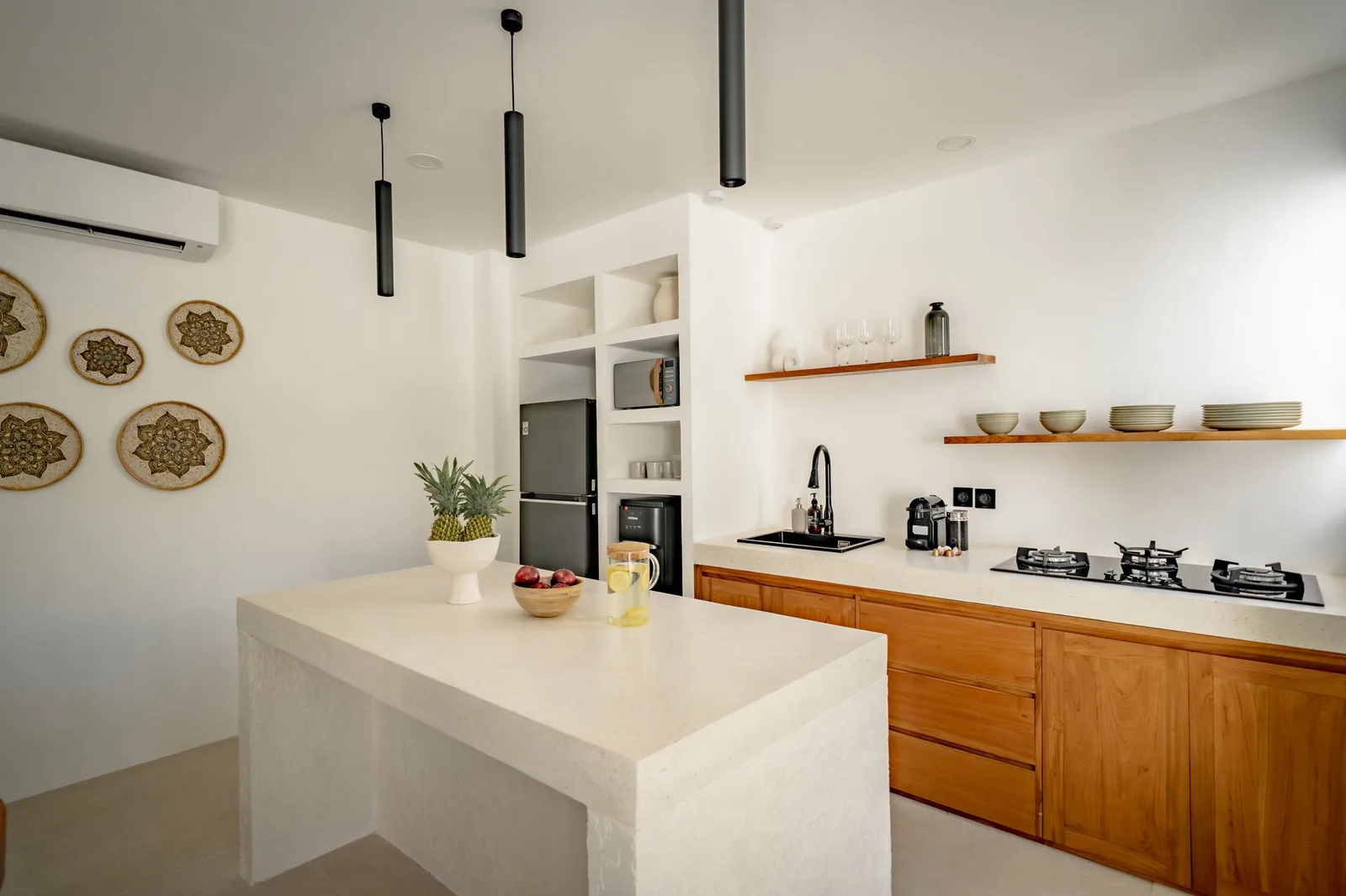 A bright modern kitchen with a large white island, wooden cabinets, open shelves, and black pendant lights.