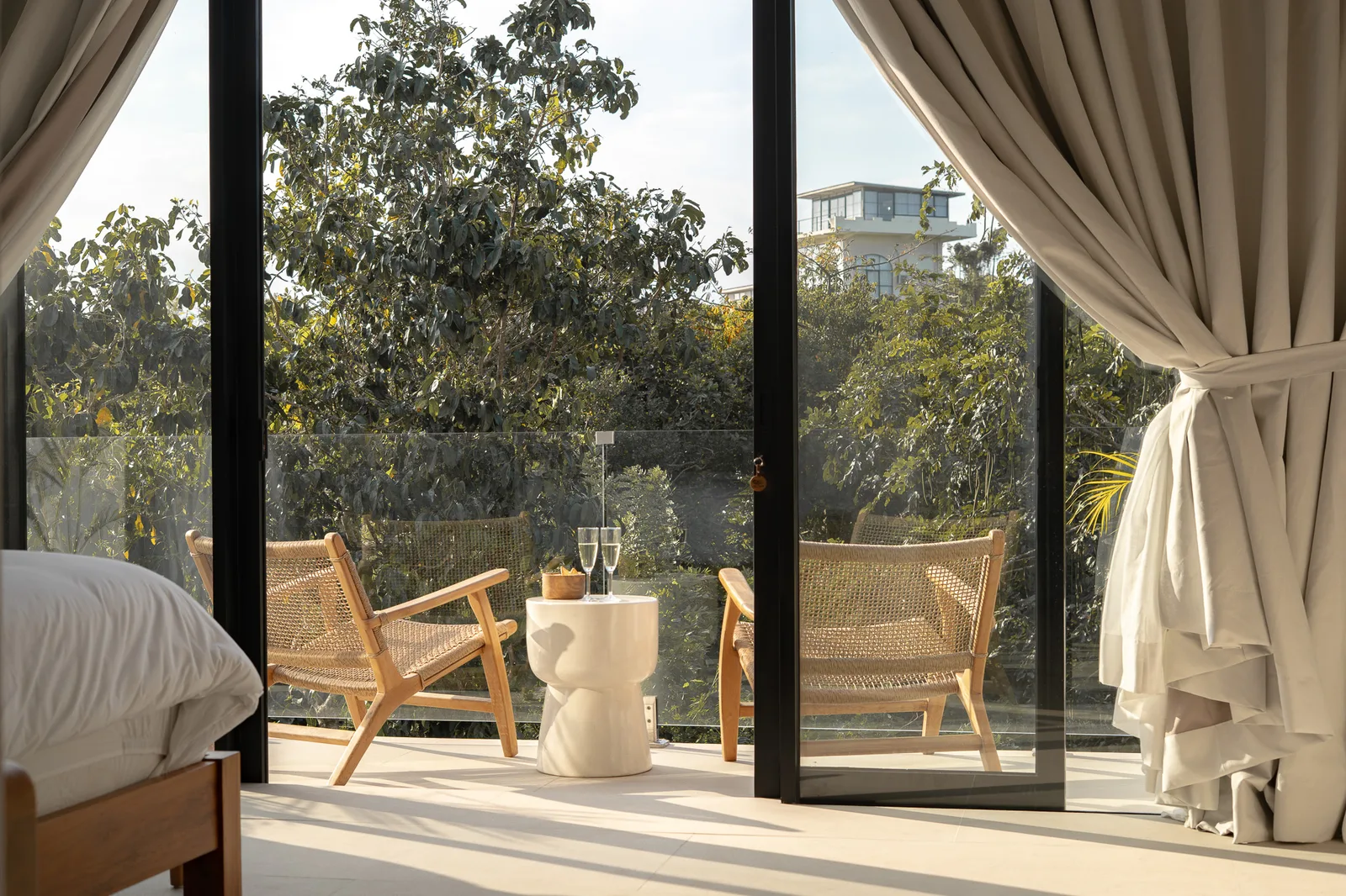 A bedroom opens onto a balcony with wicker chairs, a small table, and leafy trees beyond the glass doors.