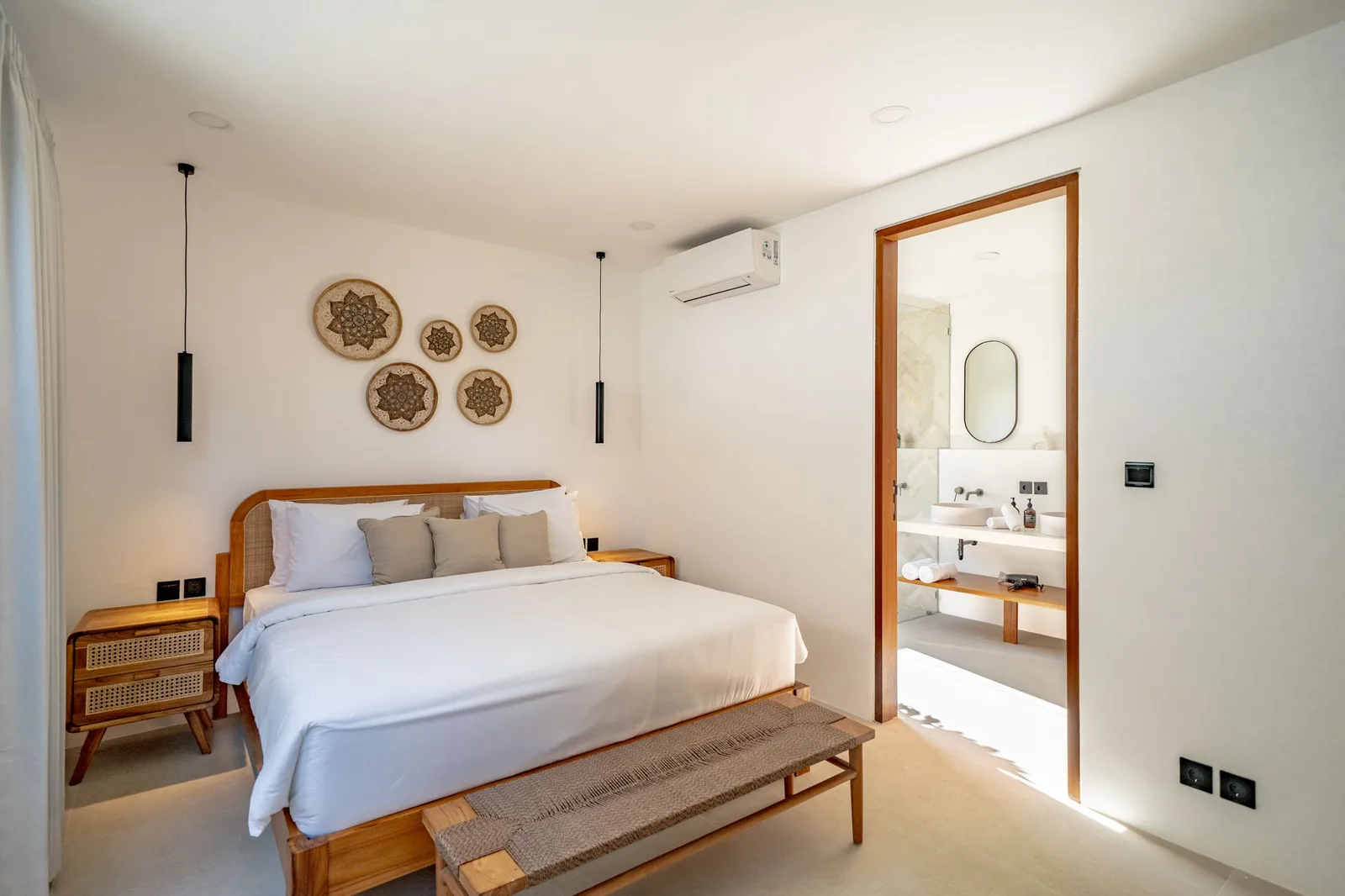 A bright bedroom has a wooden bed, woven wall decorations, pendant lights, and an open doorway to the bathroom.