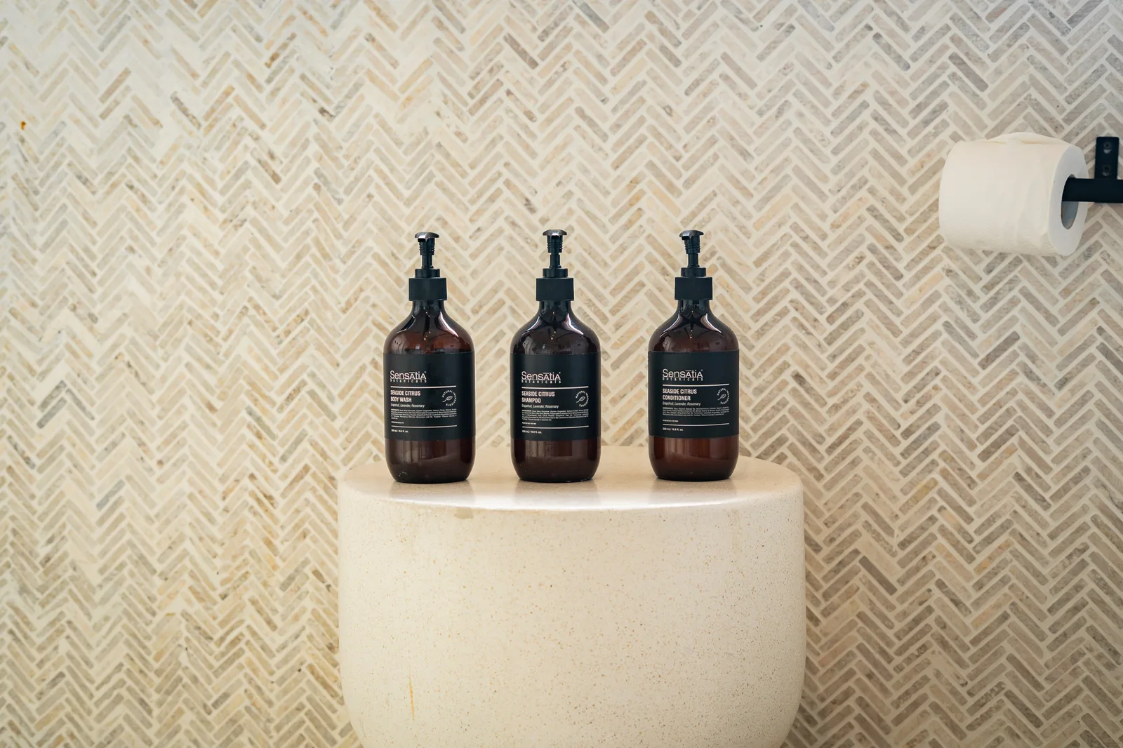 A bathroom sink holds three dark pump bottles against a patterned tile wall, with a toilet paper roll nearby.