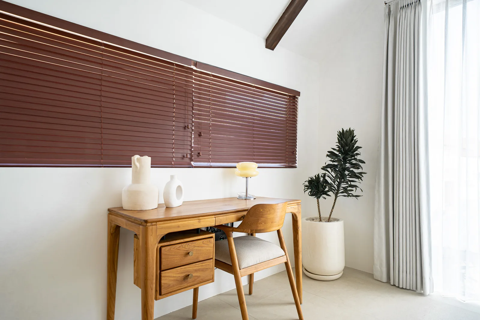 A wooden desk and chair sit beneath brown blinds, beside a potted plant and bright curtained window.