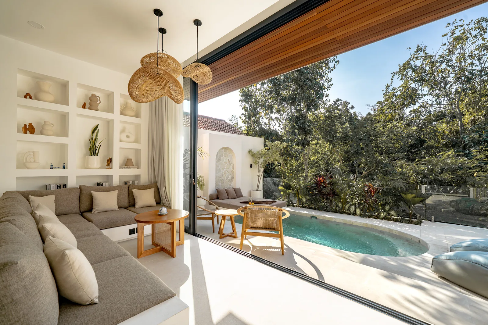 A bright living room opens onto a poolside patio, with built-in shelves, a sectional sofa, and woven pendant lights.