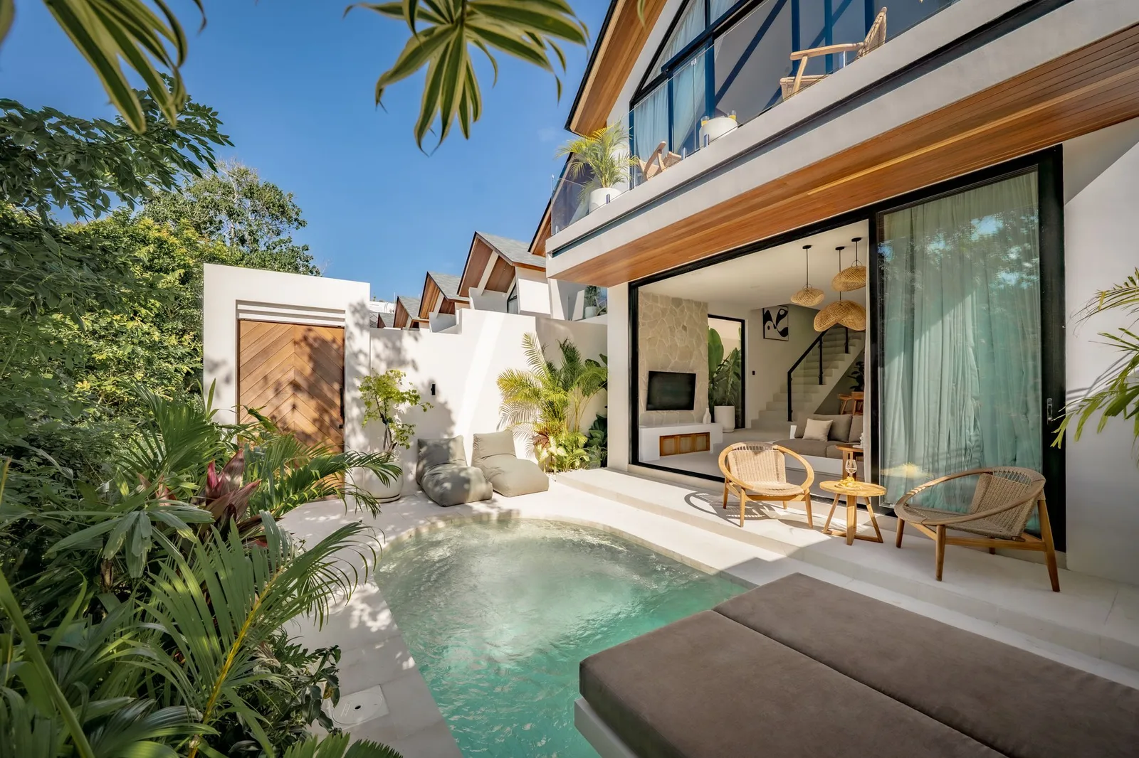 A modern villa opens onto a small turquoise pool, with a wood deck, outdoor chairs, and lush tropical plants.
