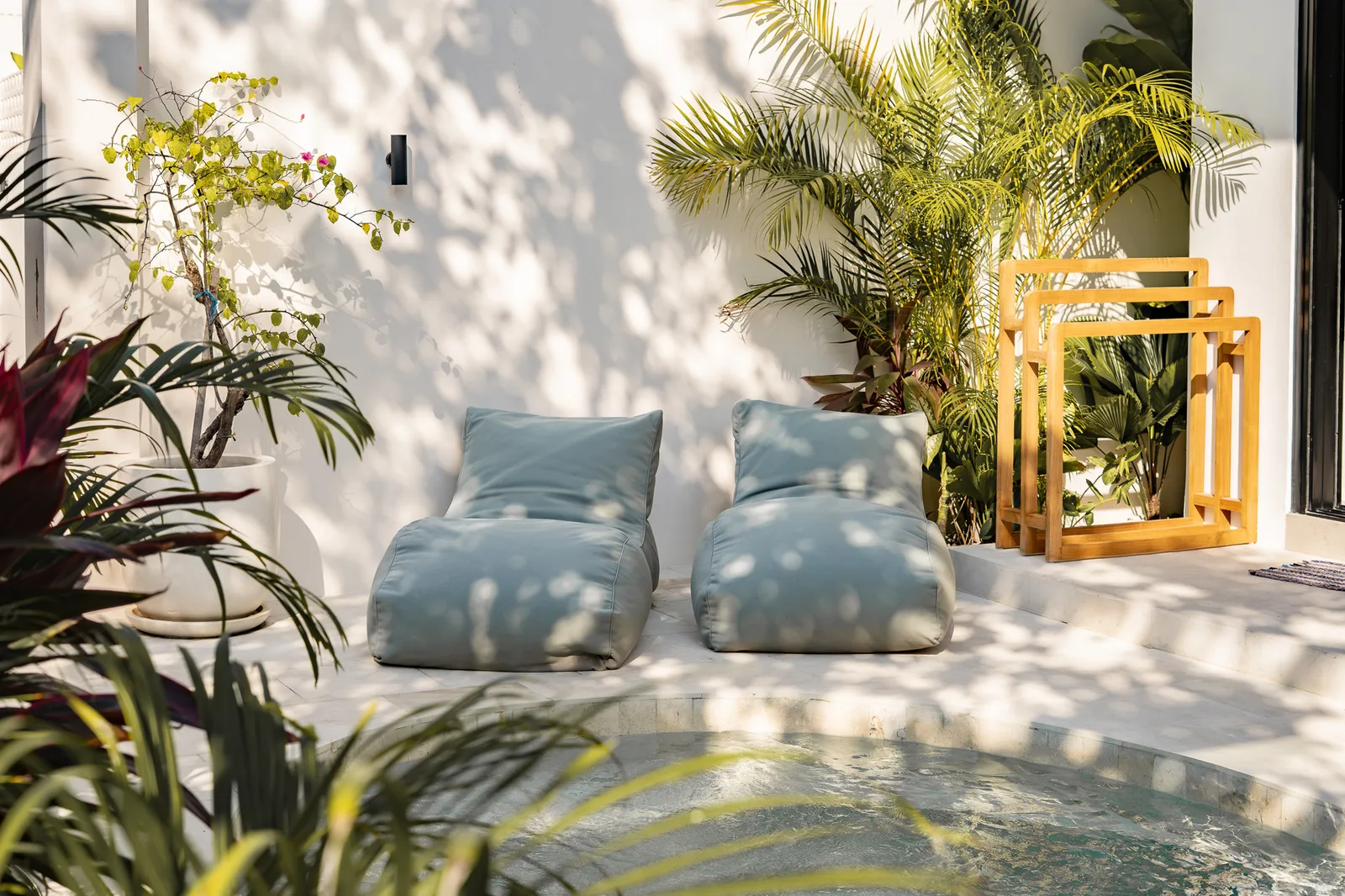 A sunny outdoor poolside seating area has two cushioned loungers, leafy plants, and wooden chairs.