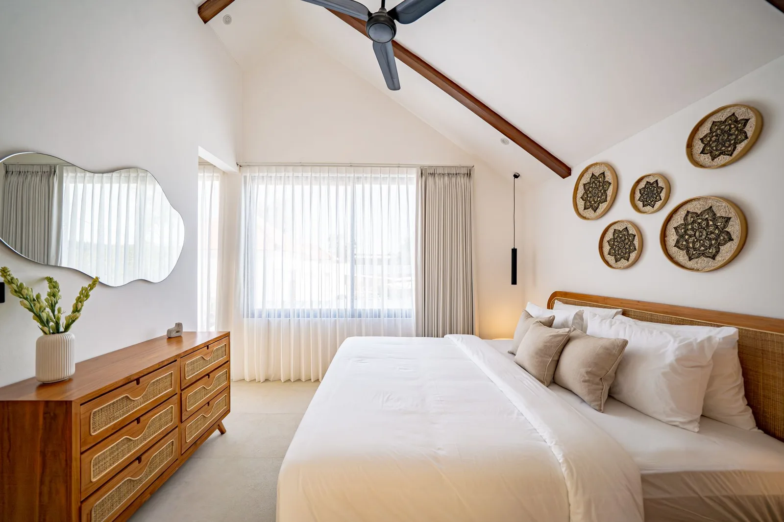 A bright bedroom has a large bed, a wooden dresser, a curtained window, and woven wall decorations.