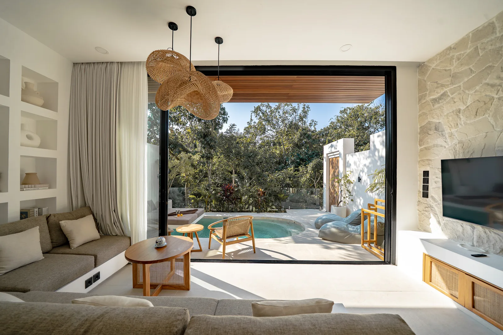 A bright living room opens onto a patio with a small pool, outdoor seating, and leafy trees.