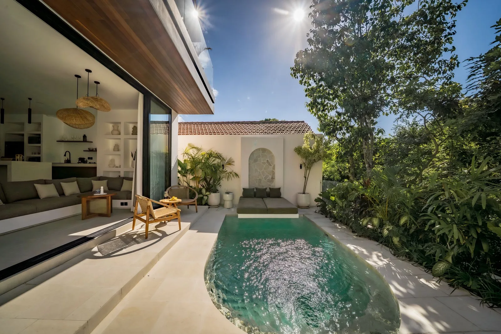 A small courtyard pool sits beside an open living area, with leafy plants and a tiled white wall.