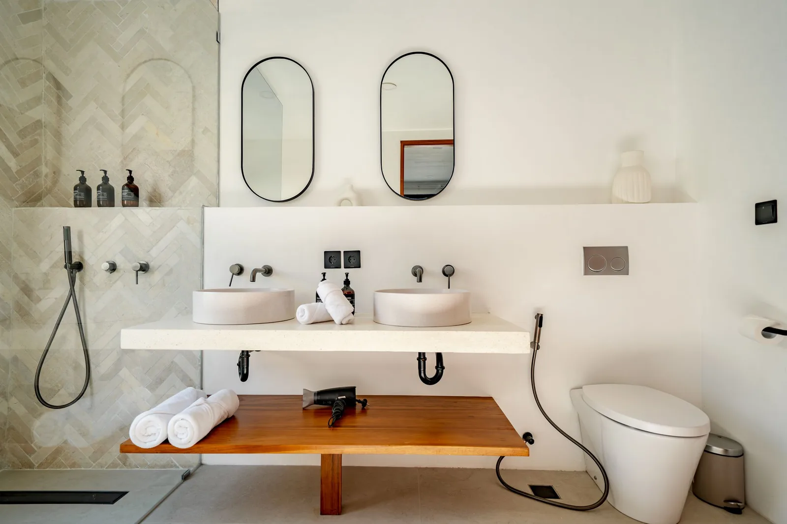 A bright modern bathroom with twin vessel sinks, arched mirrors, a wooden counter, and a freestanding bathtub.