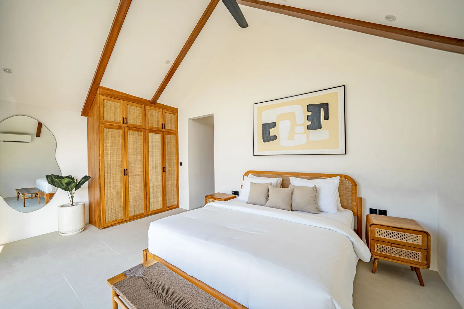 A bright bedroom with a white bed, woven rattan wardrobe and matching bedside table beneath exposed wooden ceiling beams.