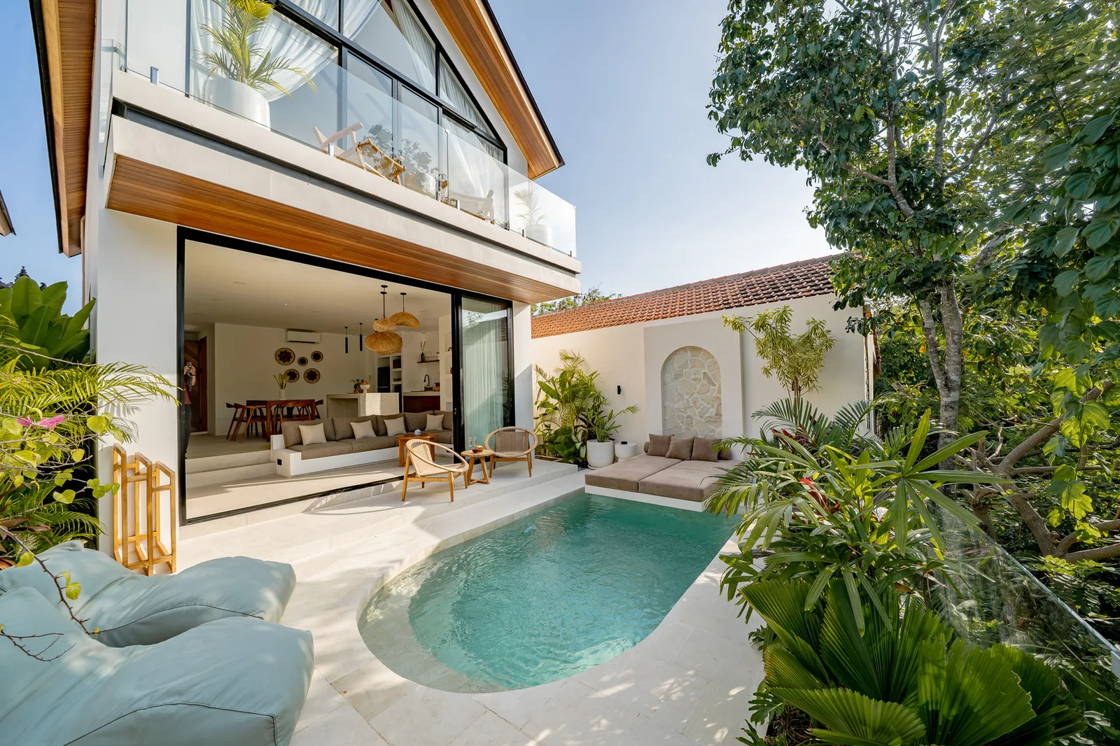 A modern house opens onto a curved swimming pool, with a furnished patio and tropical plants.