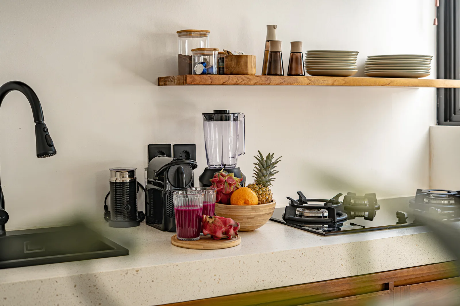 A bright kitchen counter holds a blender, coffee maker, fruit bowl, and built-in cooktop beneath an open shelf.
