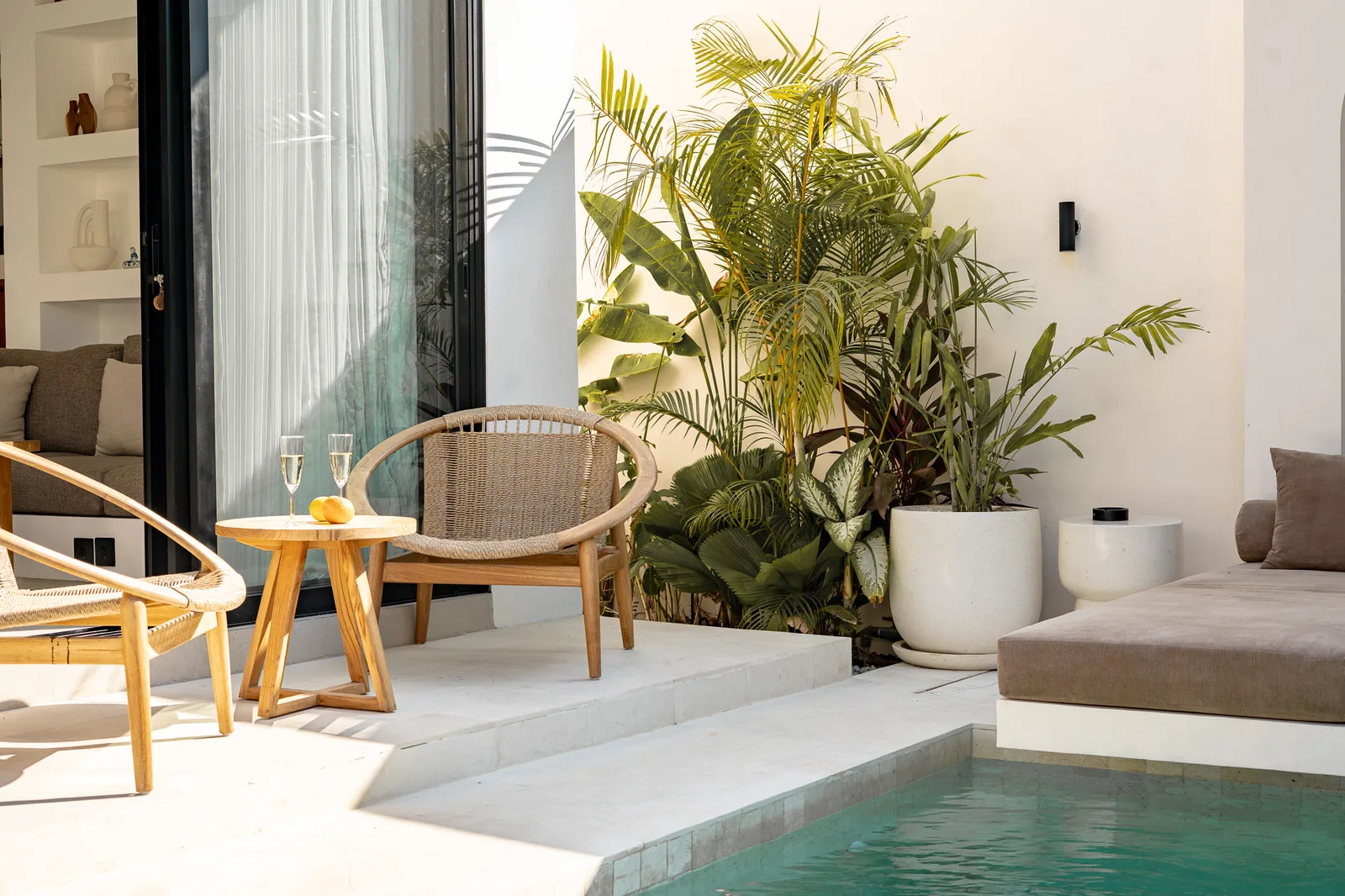 A sunlit patio has a small seating area, potted palms, and a swimming pool along the edge.