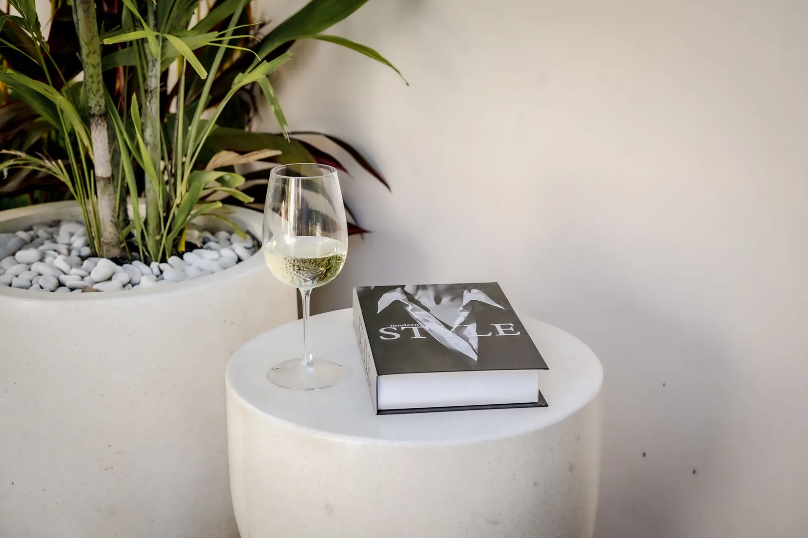 A glass of white wine and a book sit on a round white table beside a leafy potted plant.